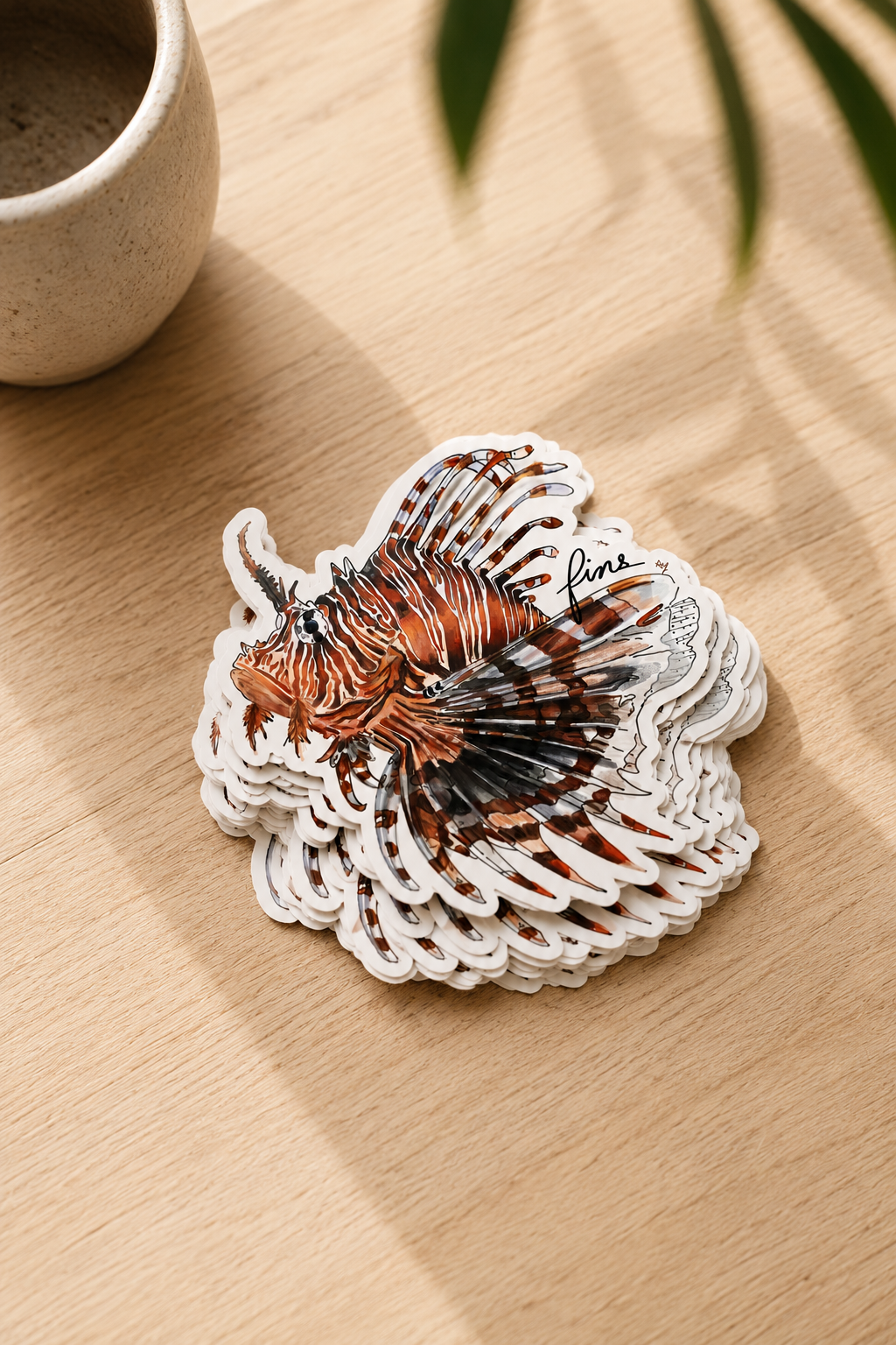 A stack of die-cut lionfish stickers on a light wood tabletop, with a ceramic cup and blurred green leaves in the background.