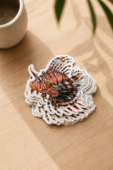 A stack of die-cut lionfish stickers on a light wood tabletop, with a ceramic cup and blurred green leaves in the background.